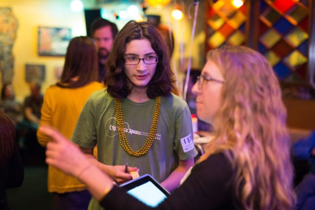Teenager with long hair wearing a green shirt and yellow beads engaging in conversation with a woman holding a tablet, at a lively event supporting My Voice Music.